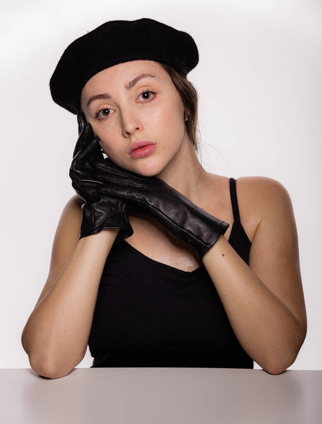 Portret van een prachtig jong modemodel in zwarte baret en leren handschoenen leunend op de tafel op een witte achtergrond. Close-up gezicht van jonge mooie vrouw met een gezonde schone huid - Foto, afbeelding