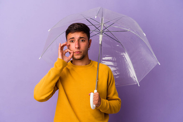 Молодой кавказский мужчина держит зонтик изолированный на фиолетовом фоне с пальцами на губах, держащий в секрете. - Фото, изображение