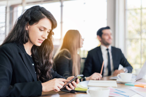 Eine kaukasische Managerin im schwarzen Anzug sitzt am Smartphone, eine Geschäftsfrau tippt auf einem Touchscreen ihres Handys, während sie sich im Büro mit verschwommenen Kollegen im Hintergrund trifft.. - Foto, Bild