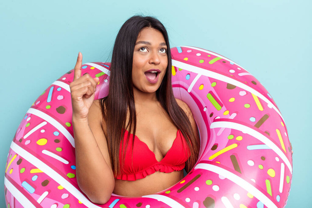 Young Venezuelan woman holding donut inflatable isolated on blue background pointing upside with opened mouth. - Photo, Image