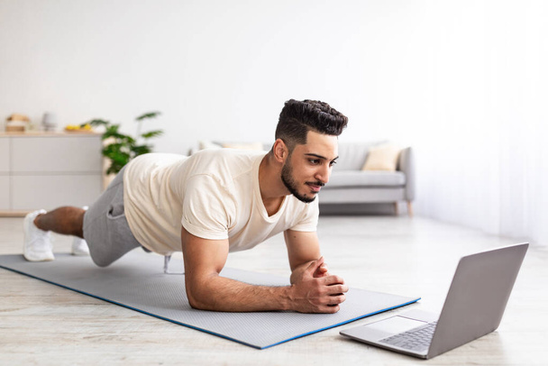 Online sporten tijdens coronavirus quarantaine. Fit jonge Arabische man doet oefeningen in de buurt laptop thuis - Foto, afbeelding