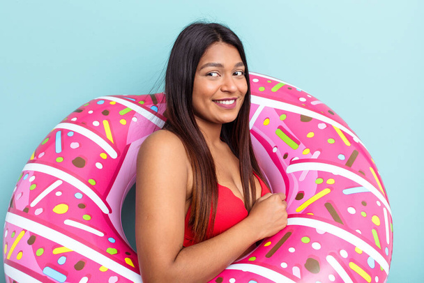 Joven venezolana sosteniendo donut inflable aislado sobre fondo azul mira a un lado sonriente, alegre y agradable. - Foto, Imagen