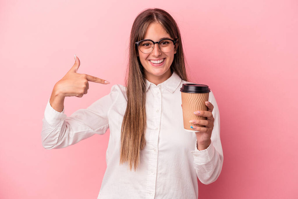 Young business caucasian woman holding a take away isolated on pink background person pointing by hand to a shirt copy space, proud and confident - Фото, изображение