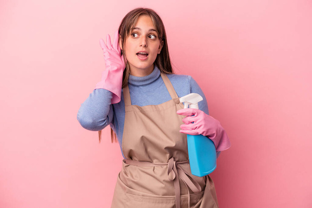 Joven mujer blanca limpiadora sosteniendo spray aislado sobre fondo rosa tratando de escuchar un chisme. - Foto, imagen