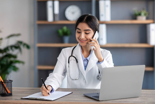 Souriant jolie jeune femme hindoue médecin en manteau blanc avec stéthoscope parler avec le patient par téléphone, travaille au bureau - Photo, image