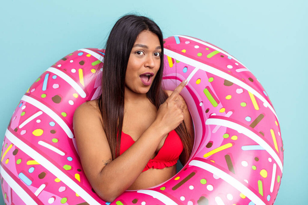 Young Venezuelan woman holding donut inflatable isolated on blue background pointing to the side - Fotó, kép