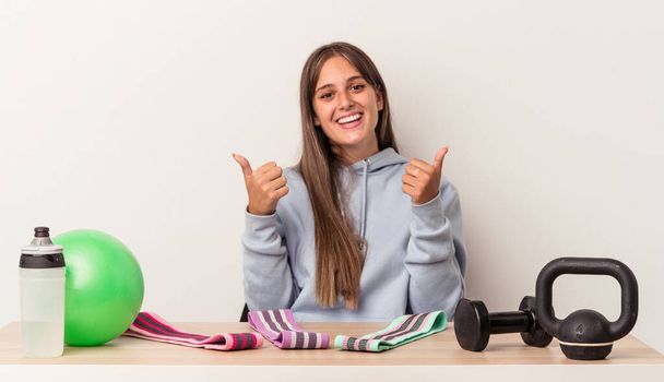 Young caucasian woman sitting at a table with sport equipment isolated on white background raising both thumbs up, smiling and confident. - Photo, Image