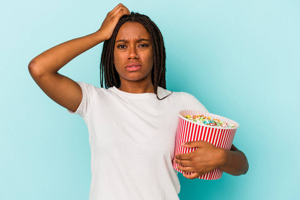 Młoda Afroamerykanka jedząca popcorny na niebieskim tle jest zszokowana, przypomniała sobie ważne spotkanie. - Zdjęcie, obraz