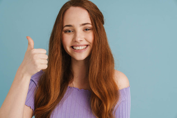Young ginger woman smiling and showing thumb up at camera isolated over blue background - Fotografie, Obrázek
