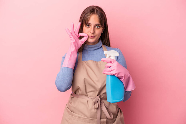 Joven mujer limpiadora caucásica sosteniendo spray aislado sobre fondo rosa con los dedos en los labios manteniendo un secreto. - Foto, Imagen