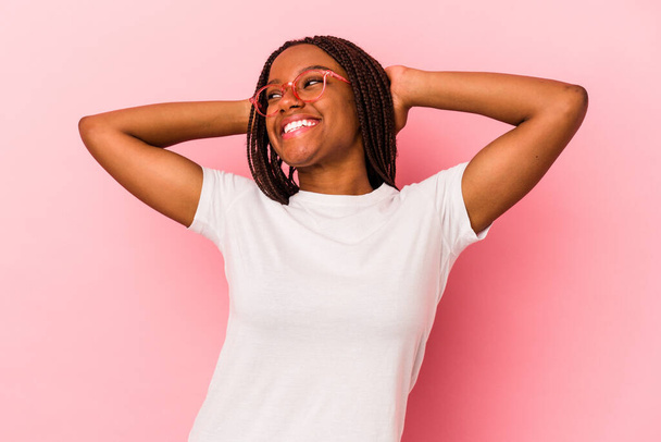 Mujer afroamericana joven aislada sobre fondo rosa sintiéndose segura, con las manos detrás de la cabeza. - Foto, imagen