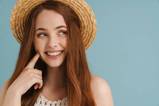 Young ginger woman in straw hat smiling and pointing finger at her cheek isolated over blue background - Foto, Imagen