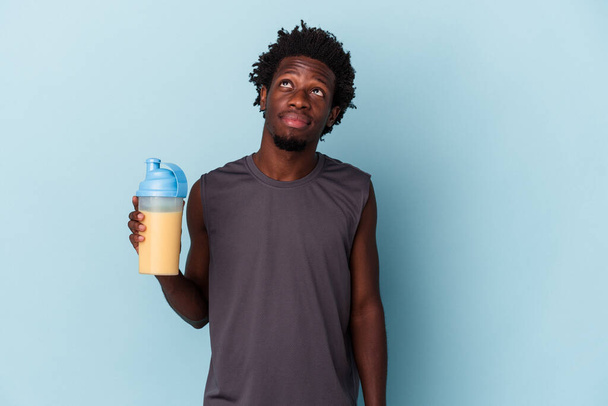Junger afrikanisch-amerikanischer Mann hält Proteinmilchshake isoliert auf blauem Hintergrund und träumt davon, Ziele und Zwecke zu erreichen - Foto, Bild