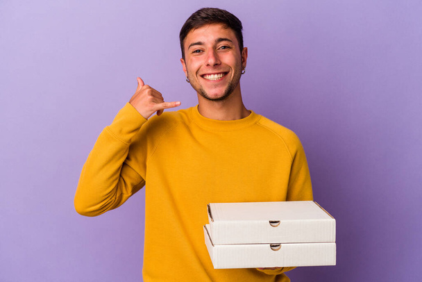 Young caucasian man holding pizzas isolated on purple background  showing a mobile phone call gesture with fingers. - 写真・画像
