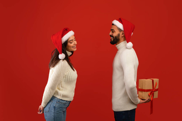 Surpresa para o Natal. Amoroso árabe homem segurando caixa de presente nas costas, parabenizando esposa sobre fundo estúdio vermelho - Foto, Imagem