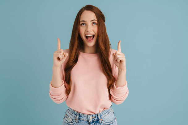 Young ginger woman smiling and pointing fingers upward isolated over blue background - Foto, Imagen