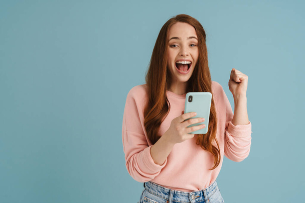 Young ginger woman making winner gesture while using mobile phone isolated over blue background - Foto, immagini