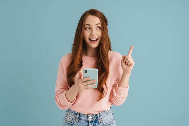 Fiatal gyömbér nő ujjal felfelé használata közben mobiltelefon elszigetelt kék háttér - Fotó, kép