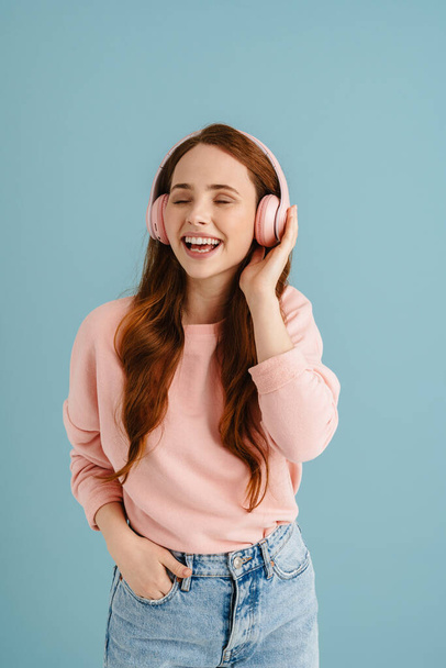 Young ginger woman laughing while listening music with headphones isolated over blue background - Fotografie, Obrázek
