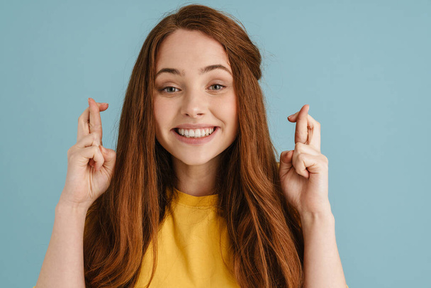 Young ginger woman smiling and holding fingers crossed for god luck isolated over blue background - Fotó, kép