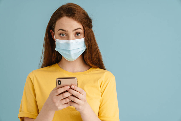 Young ginger woman in face mask using cellphone and looking at camera isolated over blue background - Fotó, kép