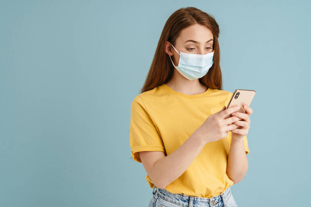Young ginger white woman in face mask using mobile phone isolated over blue background - Foto, afbeelding