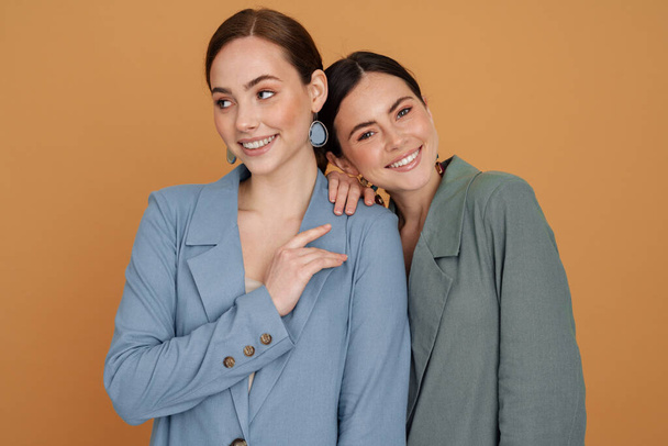 Two smiling elegant women wearing smart jackets posing standing close together isolated over beige background - Фото, изображение