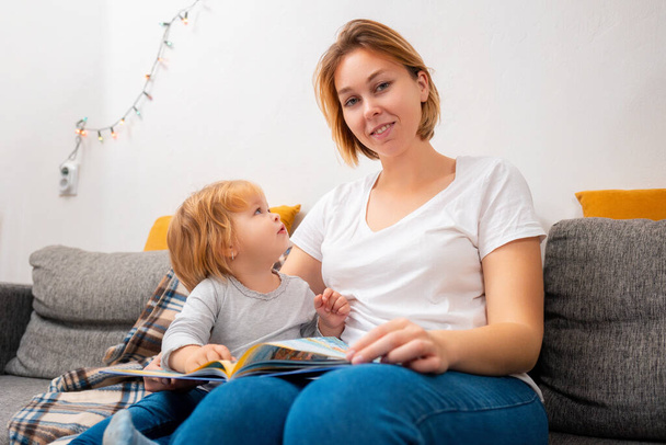 Portrait of a mother and child sitting on the sofa and reading a book. The concept of home preschool education. - Photo, Image
