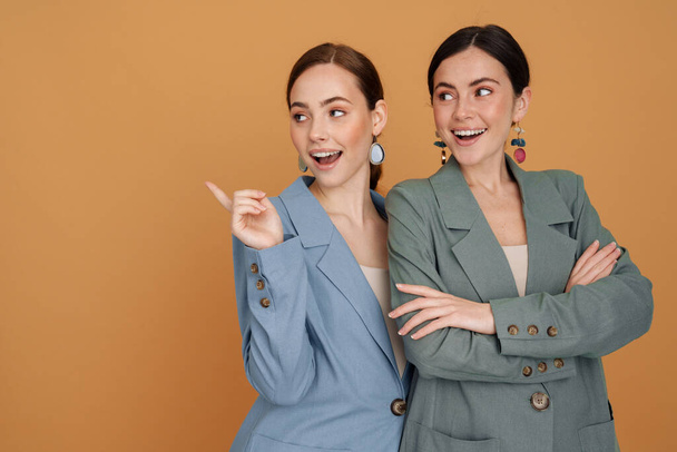 Two smiling elegant women wearing smart jackets posing standing close together isolated over beige background - Φωτογραφία, εικόνα