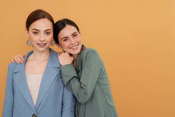 Young two women wearing jackets hugging and smiling at camera isolated over yellow background - Fotoğraf, Görsel