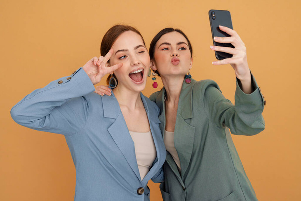 Jovens duas mulheres gesticulando ao tomar selfie no celular isolado sobre fundo amarelo - Foto, Imagem