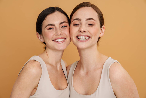 Two smiling happy young white women in underwear standing close together over beige background looking at camera showing teeth - Foto, imagen