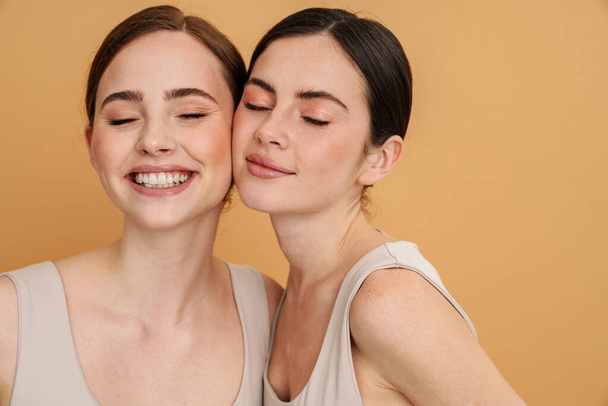 Young two women in undershirt posing and smiling at camera isolated over yellow background - Фото, зображення