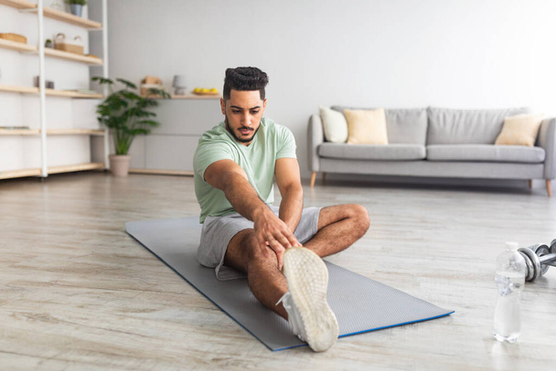Sporty young Arab guy sitting on mat, stretching leg at home, full length portrait. Copy space - Photo, Image