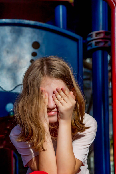 Beautiful emotional cheerful teenager girl covered her eyes with her hands on the children's city playground on a sunny day - 写真・画像