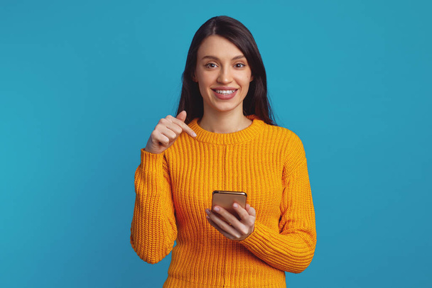 Νεαρό χαριτωμένο κορίτσι που δείχνει το smartphone, ενώ χαμογελά σε μπλε φόντο - Φωτογραφία, εικόνα