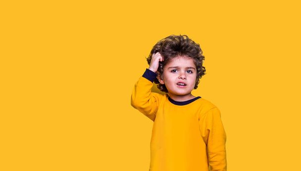 Smart funny curly kid scratches his head while thinking. Little wunderkind ready to go to school isolated on yellow background. Child looking up while thinking - Fotó, kép