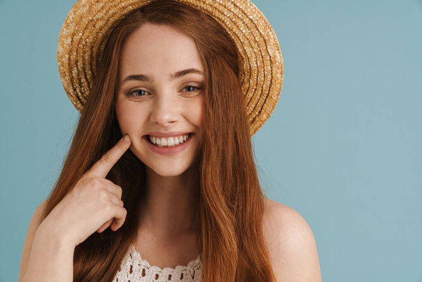 Young ginger woman in straw hat smiling and pointing finger at her cheek isolated over blue background - Foto, afbeelding