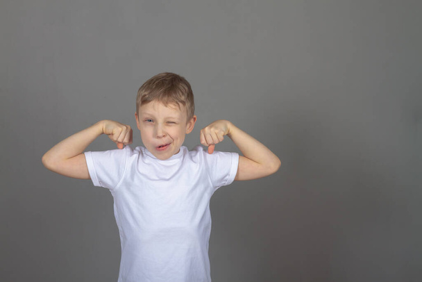 A Caucasian boy in a white T-shirt tensed his muscles on his arms, a child demonstrates his strength on a gray background in the studio. - Фото, изображение