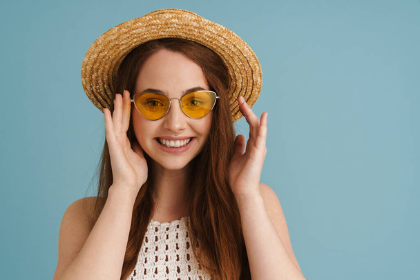 Young ginger woman in sunglasses smiling and looking at camera isolated over blue background - 写真・画像