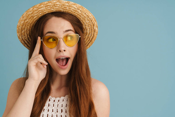 Young ginger woman in sunglasses expressing surprise and looking aside isolated over blue background - Foto, imagen
