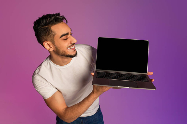 Millennial Arab man showing laptop computer with empty black screen in neon light, mockup for new website - Photo, Image