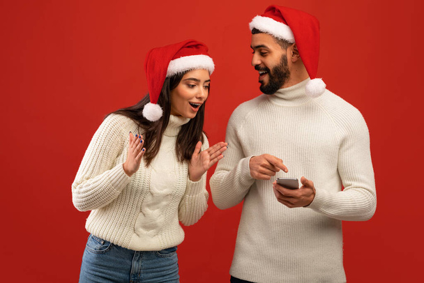 Glücklicher arabischer Mann erhält Nachricht mit Smartphone an überraschte Frau, trägt Nikolausmütze, roter Studiohintergrund - Foto, Bild