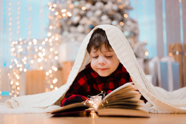 Um rapaz de pijama lê um livro debaixo de uma árvore de Natal. O humor de Ano Novo. A ler livros. Livros infantis. Um presente para o ano novo - Foto, Imagem