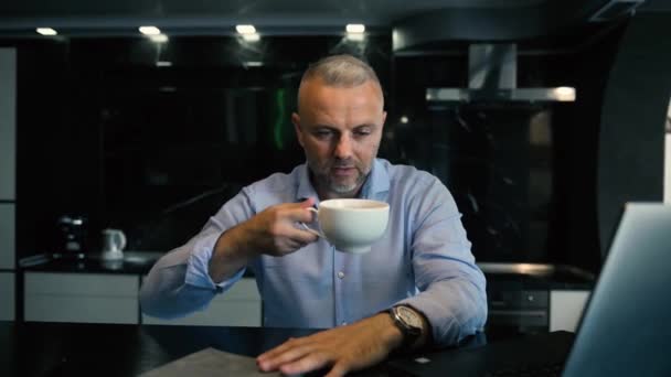 Bello uomo d'affari di mezza età di successo, imprenditore, uomo caucasico seduto di fronte a un computer portatile in cucina e godersi il caffè appena fatto mattina - Filmati, video