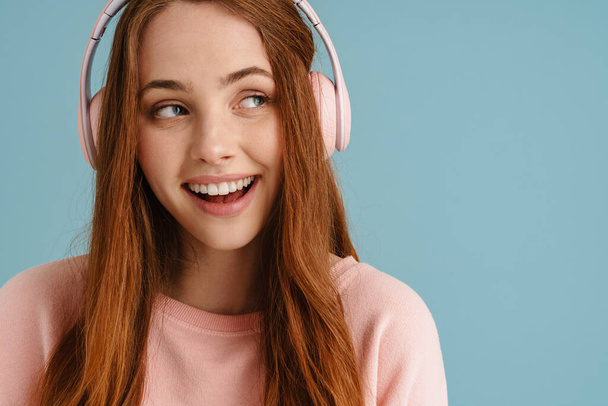 Young ginger woman smiling while listening music with headphones isolated over blue background - Φωτογραφία, εικόνα