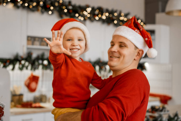 Merry Christmas and Happy New Year concept. Happy father hold his cute smiling son looking into camera and showing five fingers of his palm in festive decorated kitchen. Both men wear red sweaters and Santa hats - Zdjęcie, obraz