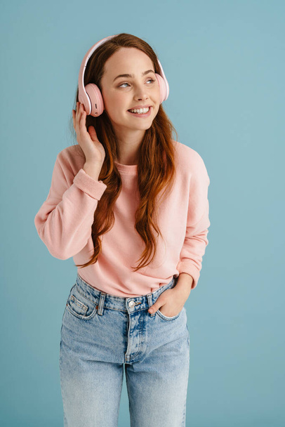 Young ginger woman smiling while listening music with headphones isolated over blue background - Fotó, kép
