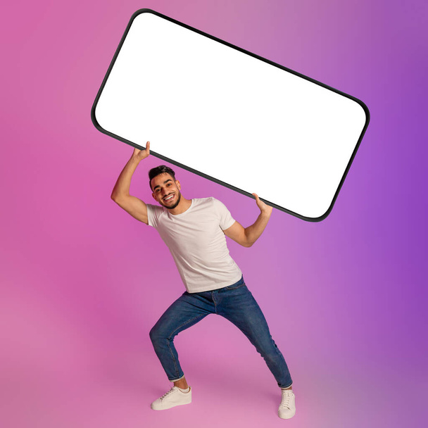 Cheerful young Arab man holding huge heavy smartphone with empty screen in neon light. Mockup - Photo, Image