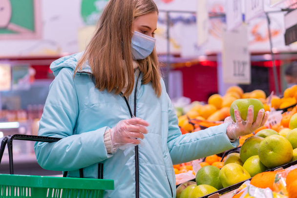 Photo of a nice young girl in a medical mask and gloves holding shopping cart choosing oranges in the supermarket. Shopping in compliance with anti-epidemic measures during the coronavirus pandemic - Photo, Image